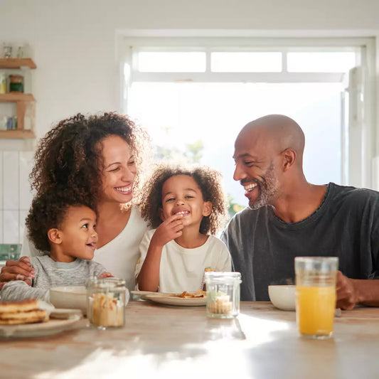 A happy family of four sitting at a breakfast table, smiling and enjoying food together in a bright kitchen with natural sunlight.